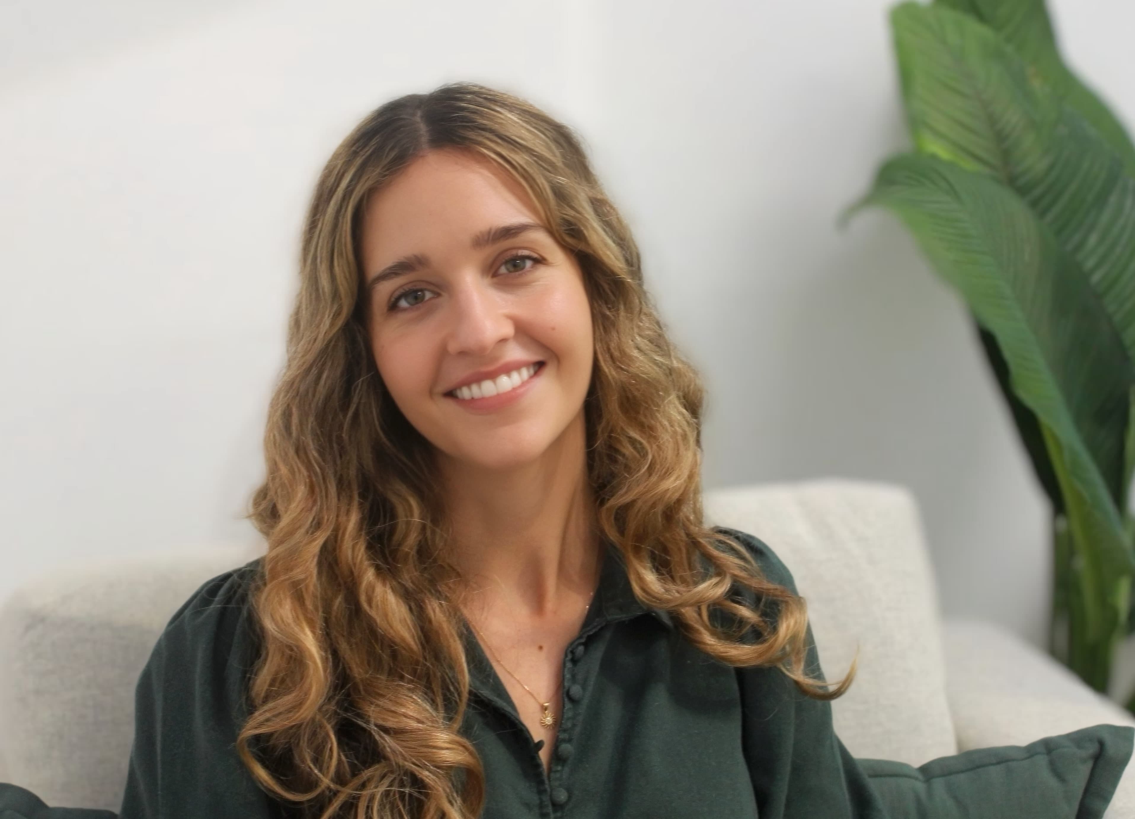 Smiling woman with long wavy blonde hair wearing a dark green blouse indoors.